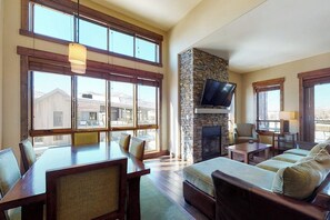 A spacious living room with a stone fireplace, large windows, and a dining area. - Spacious & impressive condo w/view, gondola access & shared hot tubs, pool, gym (Steamboat Springs)
