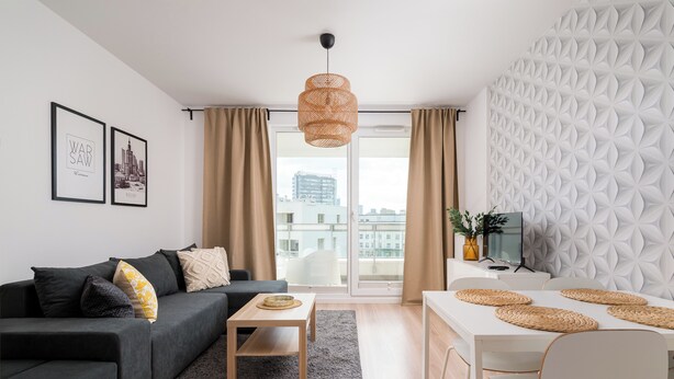 A modern living room with a sofa, coffee table, dining table with placemats, a hanging light fixture, and a patterned wall. - Kolska Chic Apartment (Warszawa)