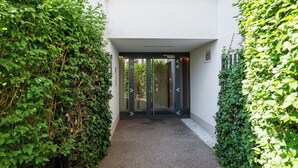 A modern building entrance with a glass door, surrounded by dense greenery. - Kolska Chic Apartment (Warszawa)