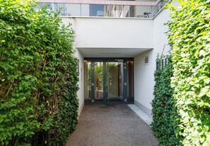 A modern building entrance with a glass door, surrounded by dense greenery. - Kolska Chic Apartment (Warszawa)