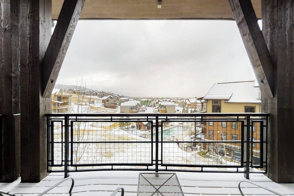A balcony with a metal railing, snow on the ground, and buildings in the background.