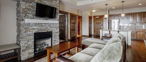 A living room with a stone fireplace, a flat-screen TV, a sofa, and a kitchen area in the background.