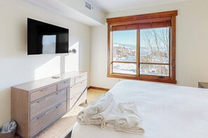 A bedroom with a bed, a TV mounted on the wall, a window with a view of a snowy landscape, and a wooden dresser. - Deluxe condo next to Wildhorse gondola w/shared outdoor pool, hot tubs, gym (Steamboat Springs)