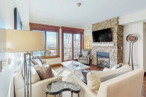 A living room with a stone fireplace, a sofa, a coffee table, and a TV mounted on the wall. - Deluxe condo next to Wildhorse gondola w/shared outdoor pool, hot tubs, gym (Steamboat Springs)
