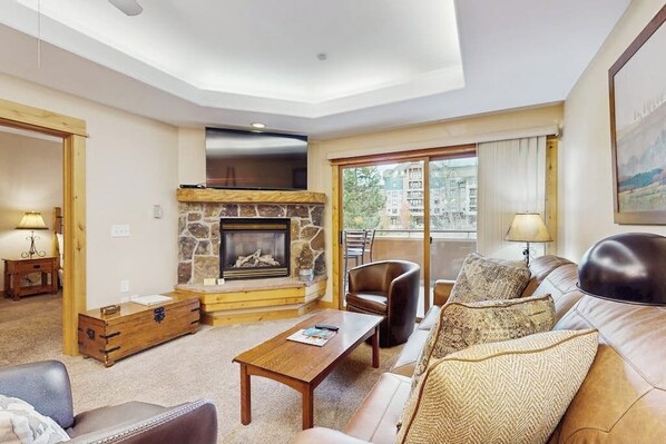 A living room with a stone fireplace, leather sofa, and a view of buildings through large windows. - Lovely, modern getaway for the whole family w/ a shared pool & gym (Steamboat Springs)