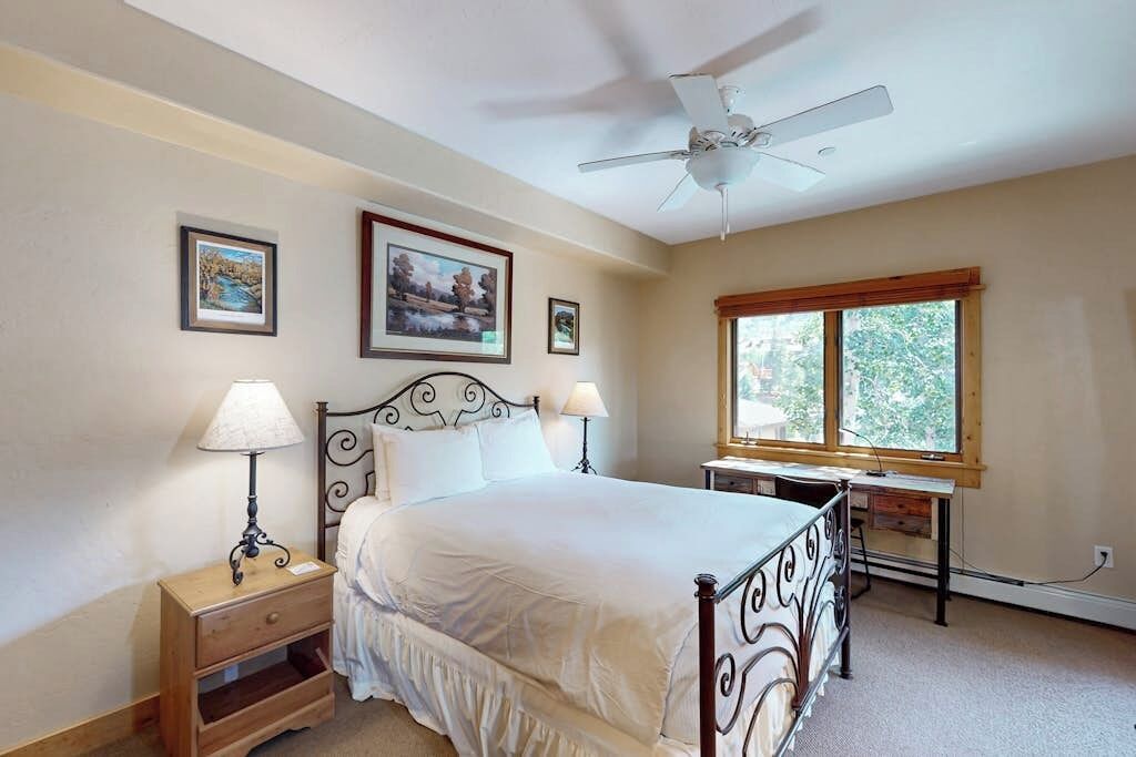 A bedroom with a bed, nightstands, a desk, a ceiling fan, and framed pictures on the wall.