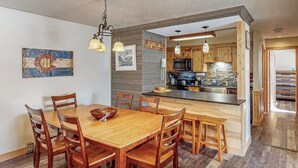 A dining area with a wooden table and chairs, a kitchen with wooden cabinets and a stone backsplash, and a sign reading 'Welcome to the Mountains.' - Deluxe condo with private patio, pool & hot tub (Steamboat Springs)