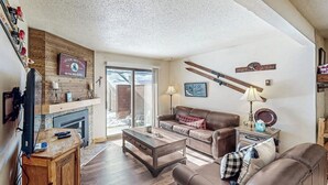 A living room with a fireplace, a brown leather sofa, a coffee table, and a television. - Deluxe condo with private patio, pool & hot tub (Steamboat Springs)