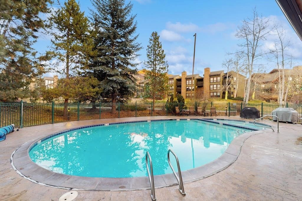 A swimming pool with a metal ladder, surrounded by a fence, trees, and a building in the background.