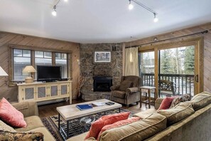 Ein gemütliches Wohnzimmer mit Kamin, Fernseher und einem Balkon mit Blick auf Bäume.