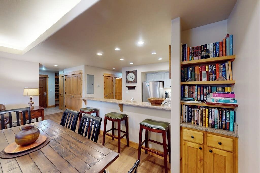 Uma cozinha espaçosa com armários de madeira, uma área de jantar com mesa e cadeiras, e uma estante repleta de livros.