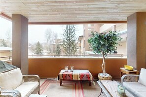 Ein Balkon mit Korbmöbeln, einem Tisch mit gemustertem Tischläufer, einem Blumentopf und Blick auf Gebäude und Bäume.