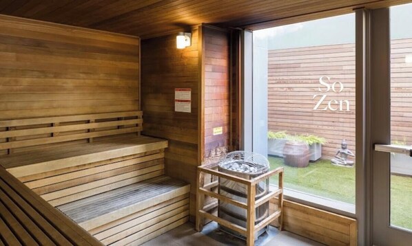 Uma sauna de madeira com bancos, uma pequena mesa com um cesto, e vista para um jardim através de uma porta de vidro.