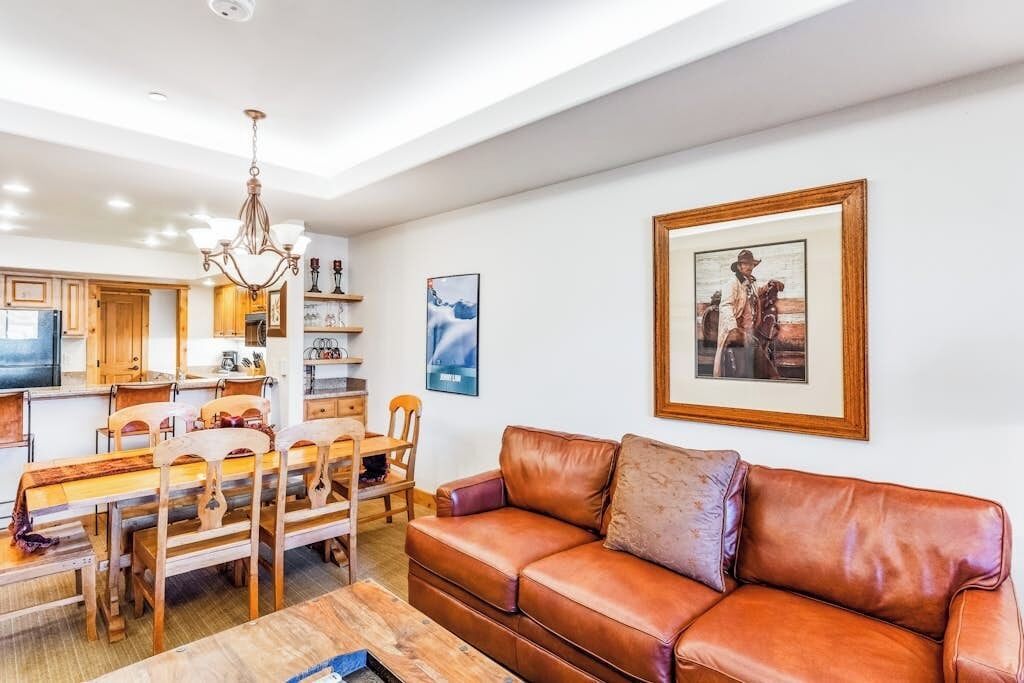 Ein Wohnzimmer mit brauner Ledersofa, einem Holzespeisetisch und Stühlen. Es gibt eine Chandelieber, ein gerahmtes Bild an der Wand und eine Küche im Hintergrund.