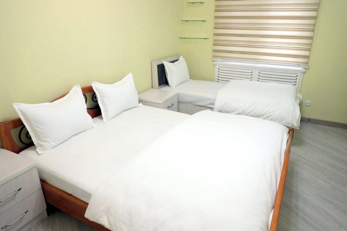 Una habitación con dos camas individuales, una mesita de noche con lámpara, un armario y una ventana con persianas.