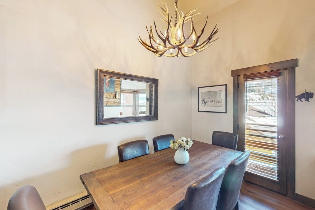 Une salle à manger avec une table en bois, des chaises, un lustre, des œuvres d’art encadrées et une fenêtre avec des stores.