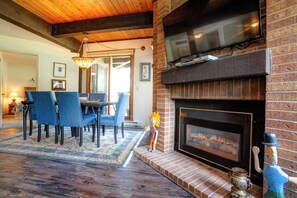 Ein Wohnzimmer mit Kamin, einem darüber montierten Fernseher und einem Essbereich mit blauen Stühlen.