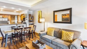 Una sala de estar con sofá, mesa de centro, mesa de comedor con sillas, área de cocina y un cuadro en la pared.