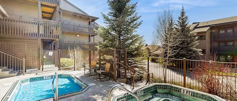 A pool area with two pools, a wooden building with balconies, and a tall evergreen tree.