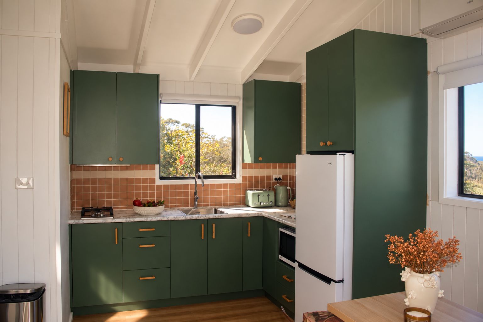 A kitchen with green cabinets, a white refrigerator, and a window with a view of trees.