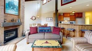 Una sala de estar con chimenea, sofá, mesa de centro y zona de cocina con armarios de madera y una barra.