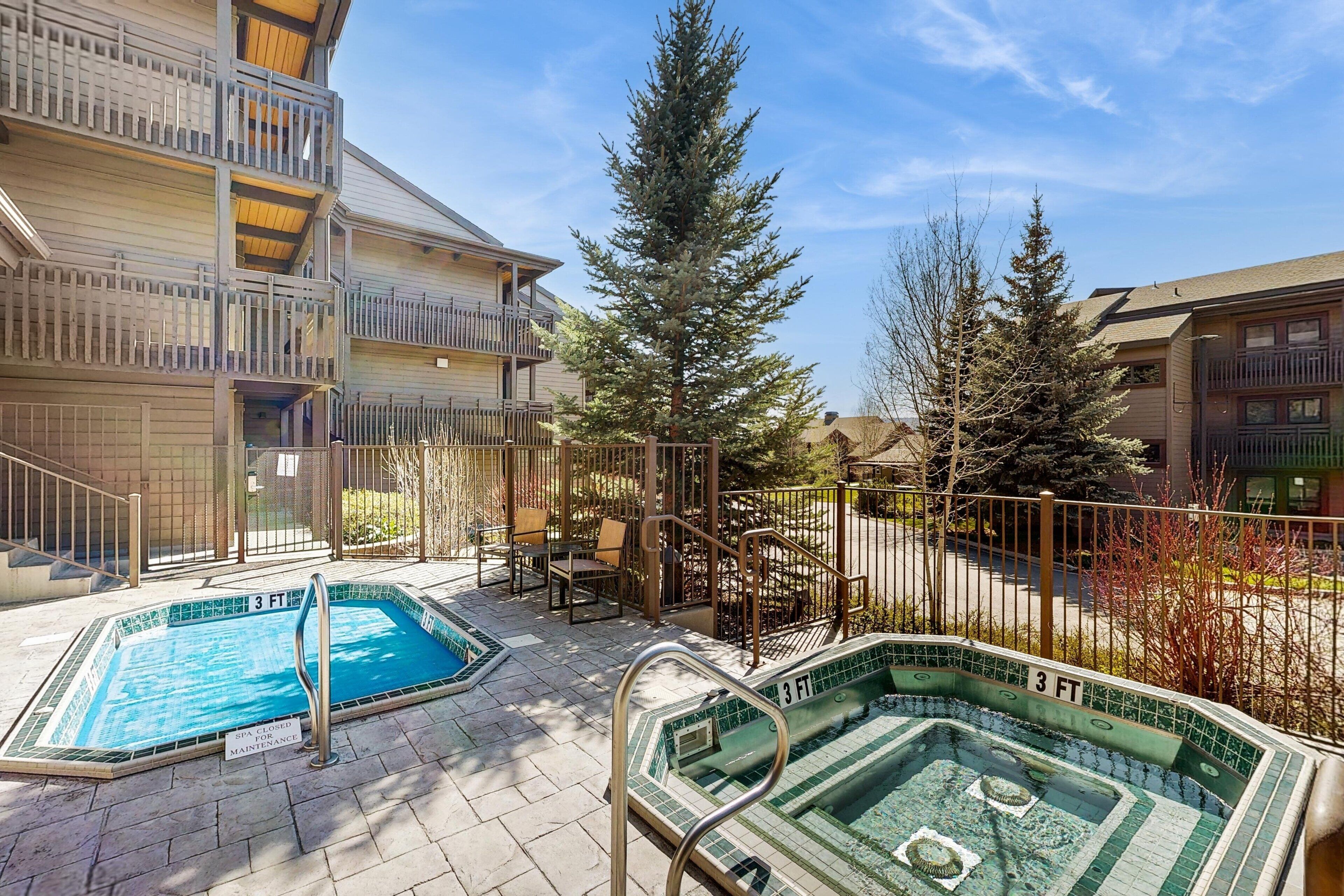 A pool area with two pools, a wooden building with balconies, and a tall evergreen tree.