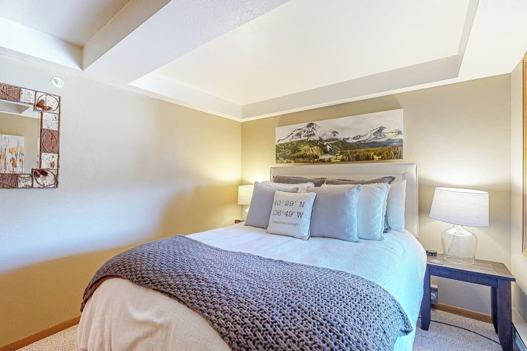 A bedroom with a bed, pillows, a nightstand, a lamp, a painting, and a decorative mirror.