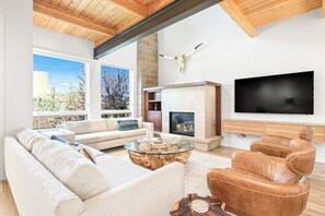 Ein modernes Wohnzimmer mit Sofa, Ledersessel, Glastisch, Kamin, Fernseher und einem großen Fenster mit Aussicht.
