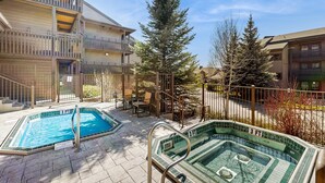Ein Poolbereich mit zwei Pools, ein Gebäude mit Balkonen und ein hoher Nadelbaum.
