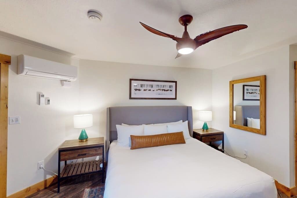 Quarto de hotel com cama, duas mesas de cabeceira, ventilador de teto, ar condicionado e um quadro na parede.