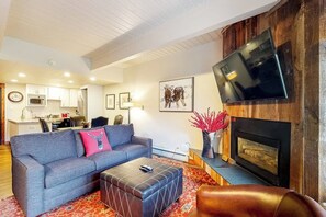 A living room with a sofa, ottoman, fireplace, TV, and kitchen in the background. - Remodeled, Ski-In/Ski-Out Studio w/ Fireplace & Shared Hot Tub - Near Hiking (Steamboat Springs)