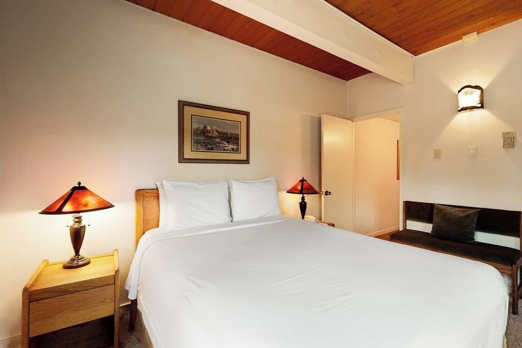Une chambre à coucher avec un grand lit, deux lampes de chevet, une table de chevet, une tête de lit en bois, un tableau encadré au mur et une porte donnant sur une autre pièce.