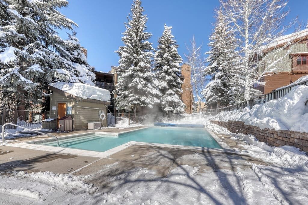 Un área de piscina con árboles cubiertos de nieve, un edificio con chimenea y un cielo azul despejado.