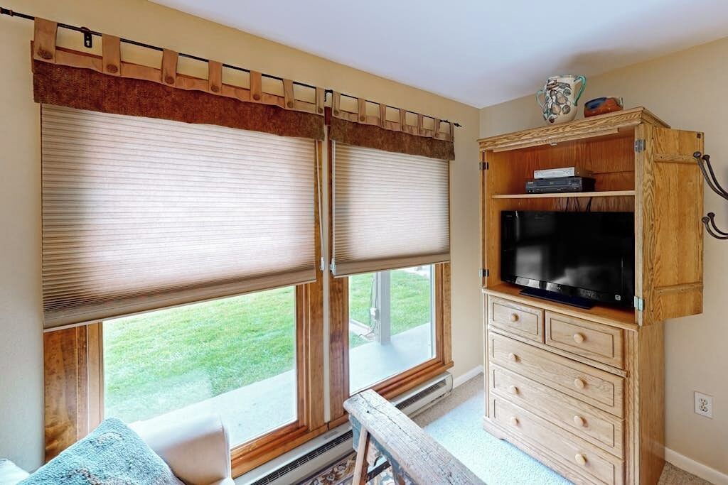 Una habitación con dos ventanas amplias con persianas, un mueble de madera con un televisor y un banco.