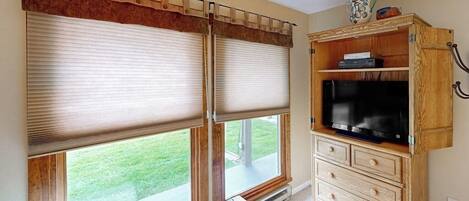 Una habitación con dos ventanas amplias con persianas, un mueble de madera con un televisor y un banco.