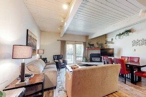Ein Wohnzimmer mit Sofa, Sessel, Esstisch und Stühlen. Ein Kamin, ein Fernseher und ein Hirschgemälde an der Wand sind ebenfalls vorhanden.