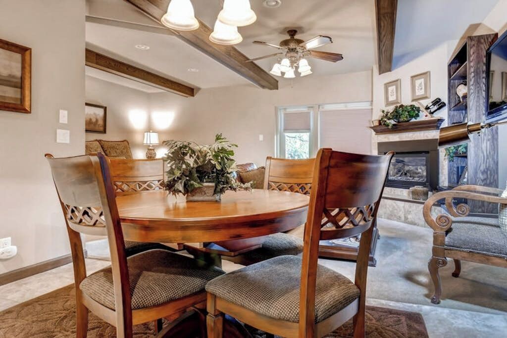 A dining area with a round wooden table, cushioned chairs, a ceiling fan, and a fireplace.