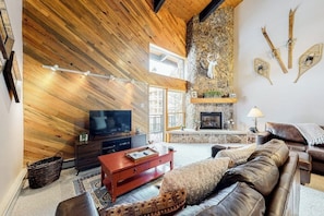 A living room with a stone fireplace, wooden ceiling, and a television. - Top-Floor, Ski-In/Ski-Out Condo w/ Loft, Fireplace, Balcony & Shared Hot Tub (Steamboat Springs)