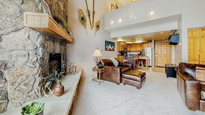 A spacious living area with a stone fireplace, leather furniture, and a kitchen in the background. - Top-Floor, Ski-In/Ski-Out Condo w/ Loft, Fireplace, Balcony & Shared Hot Tub (Steamboat Springs)