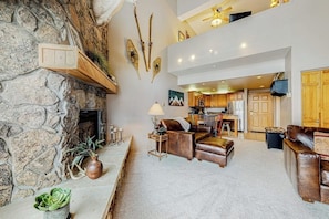 A spacious living area with a stone fireplace, leather furniture, and a kitchen in the background. - Top-Floor, Ski-In/Ski-Out Condo w/ Loft, Fireplace, Balcony & Shared Hot Tub (Steamboat Springs)