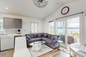 Un salon moderne avec un canapé d’angle gris, une table basse en verre, un fauteuil blanc et un coin repas comprenant une table et des chaises.