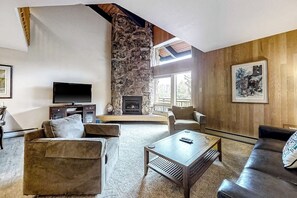 A living room with a stone fireplace, a television, and a large window. - Lofty Ski-In/Ski-Out Condo w/ Fireplace, Balcony & Shared Hot Tub/Grill/Laundry (Steamboat Springs)