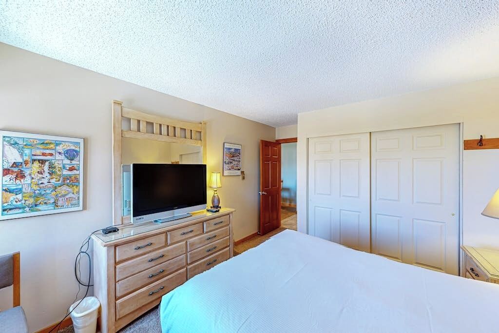 A bedroom with a bed, a TV on a wooden dresser, a lamp, a painting, and a wardrobe.