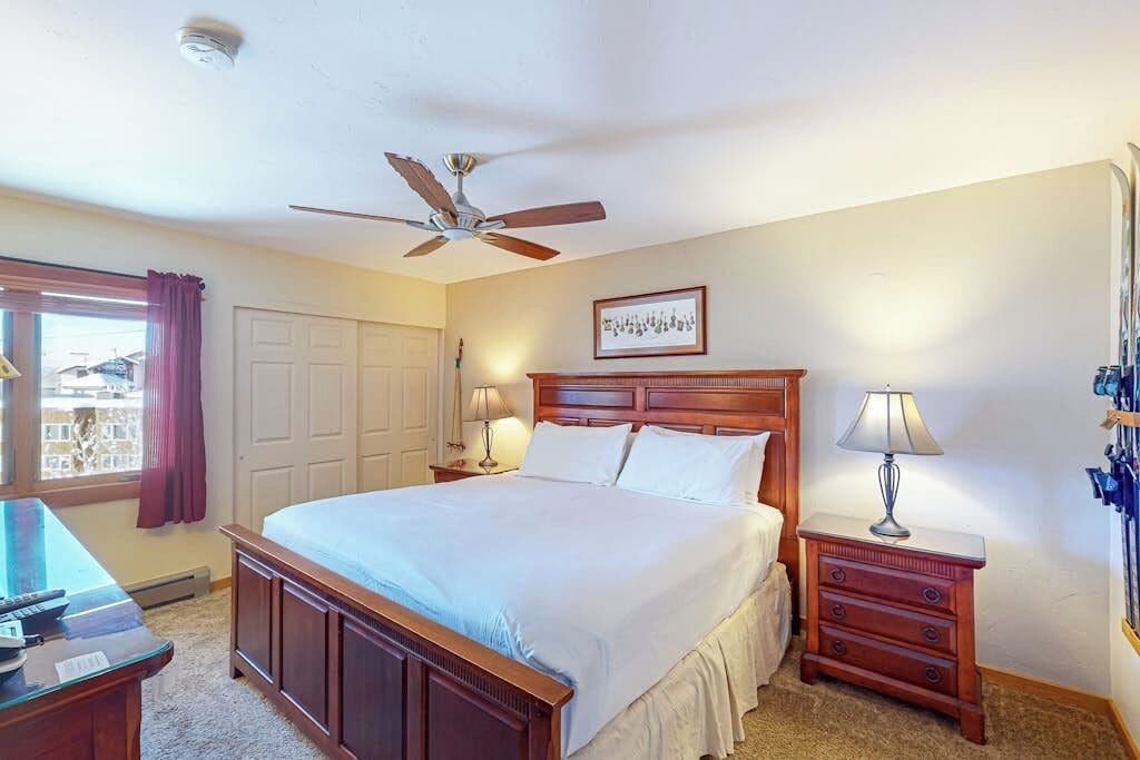 A bedroom with a bed, a desk, a chair, a ceiling fan, and a window with curtains.