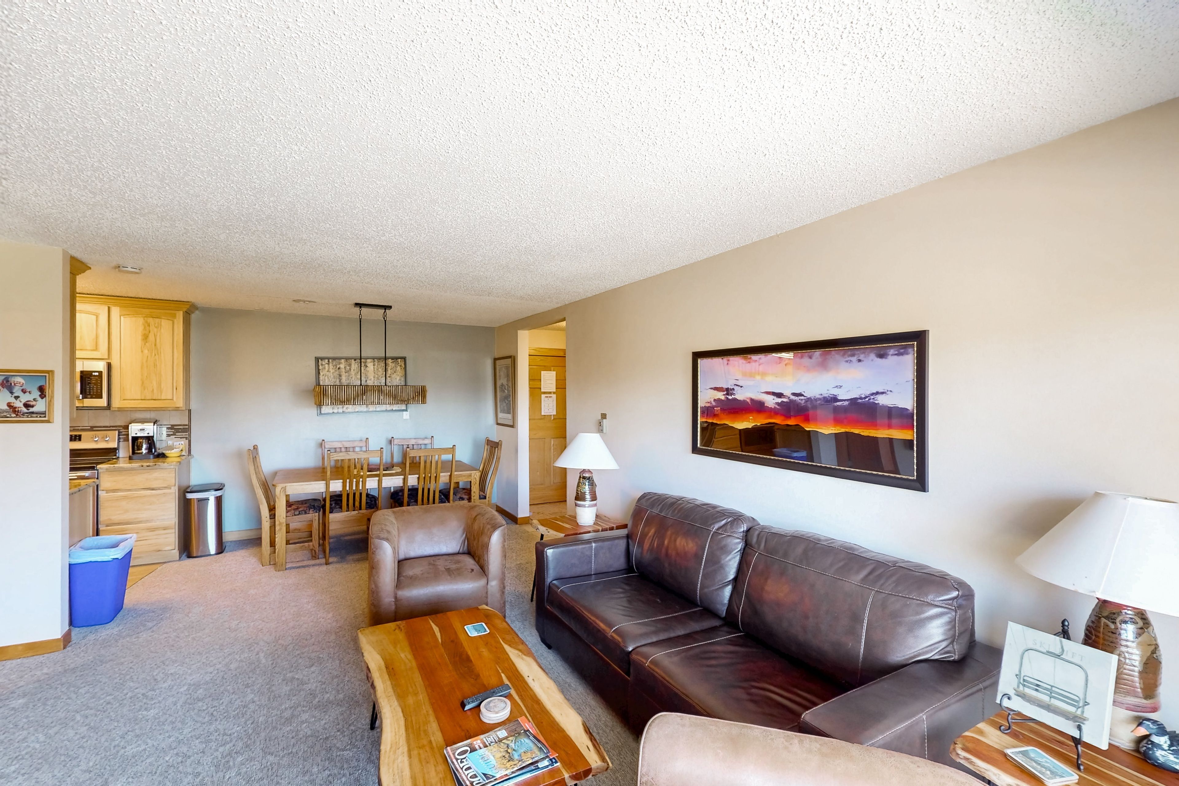 A living room with a brown leather sofa, a wooden coffee table, and a painting of a sunset.