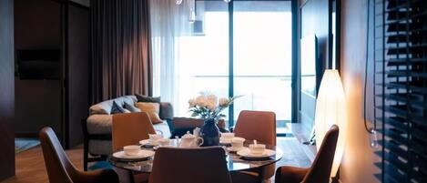 Una habitación de hotel moderna con zona de comedor, sofá, televisor y un ventanal grande.