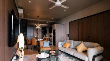 A modern living room with a sofa, coffee table, and TV. There is a dining area with chairs and a table. The room has a ceiling fan and recessed lighting.