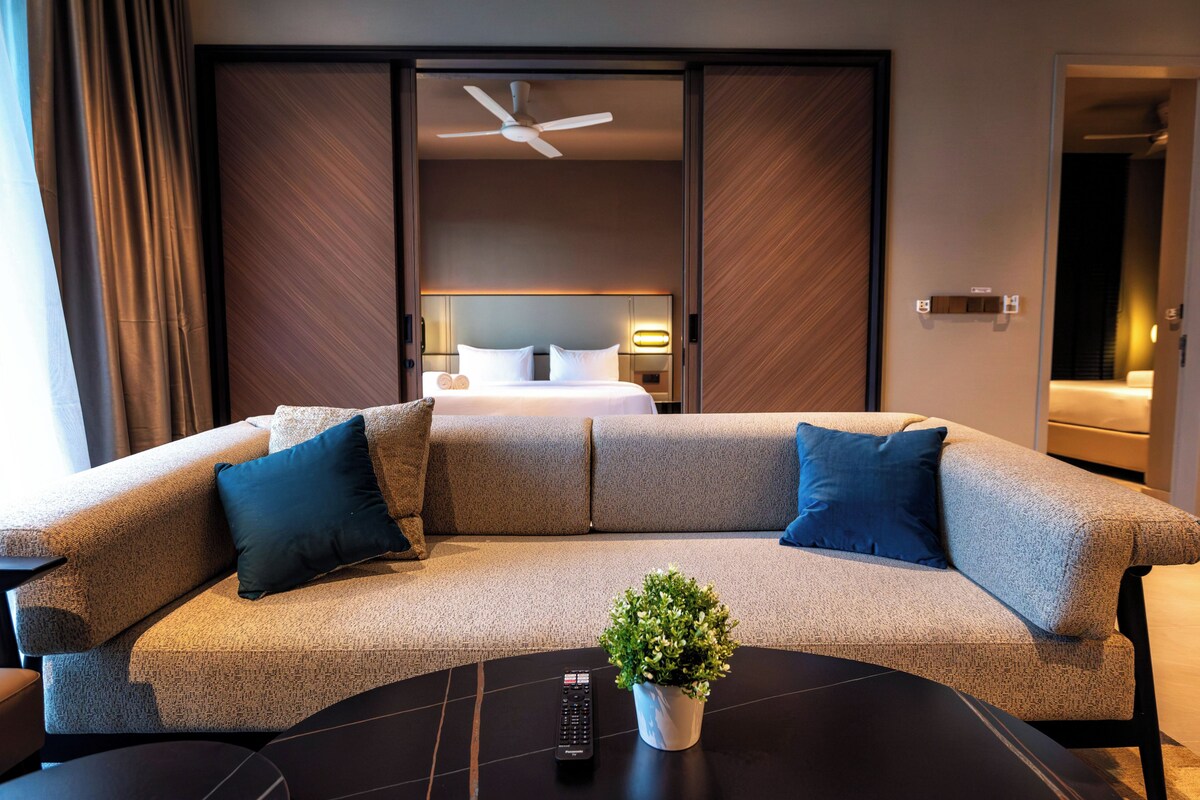 Habitación de hotel moderna con un sofá curvo, una mesita con una planta y una cama con ropa de cama blanca.