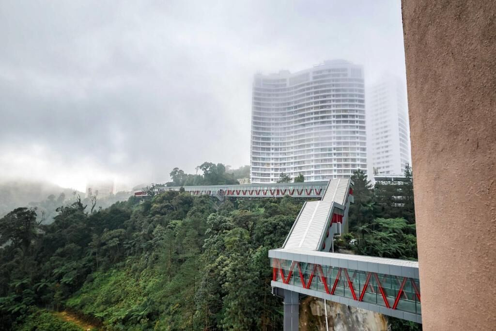 Un edificio moderno con fachada de vidrio, un puente peatonal con barandillas rojas y una ladera verde y exuberante.