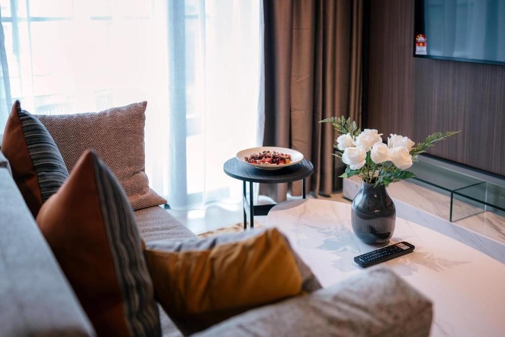 A modern living room with a sofa, a small round table with a bowl of fruit, a vase with white flowers, and a remote control.